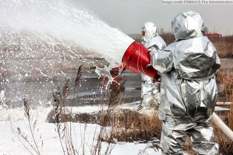 Two people in silver hazmat suits spraying a foam solution into a body of water