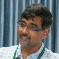 Indian man wearing glasses and a green/blue shirt standing against a blue curtain
