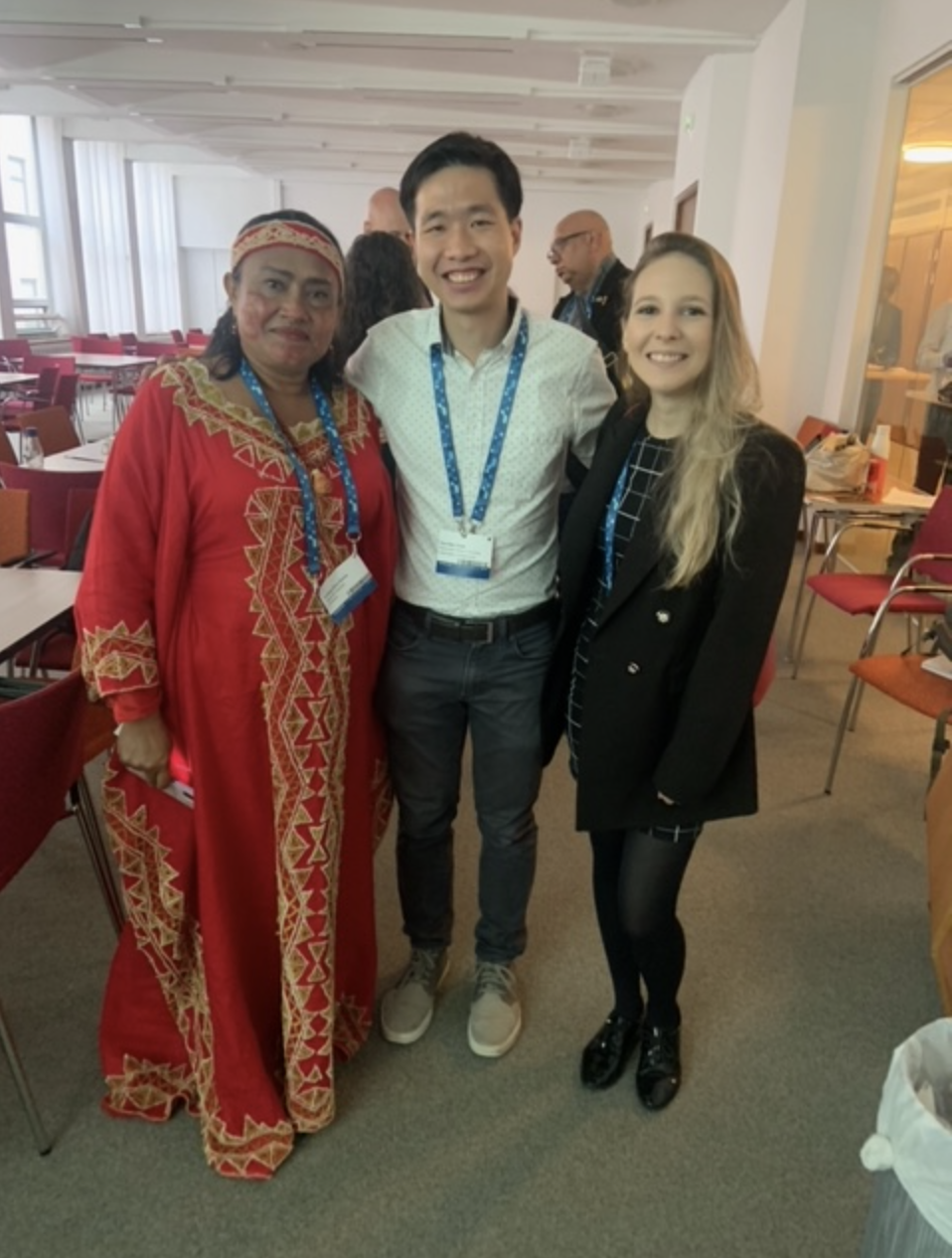 A man standing between two women. Woman on the left wears a long red dress. Man in middle wears a whit shirt and gray pants. Woman on the right wears a black dress