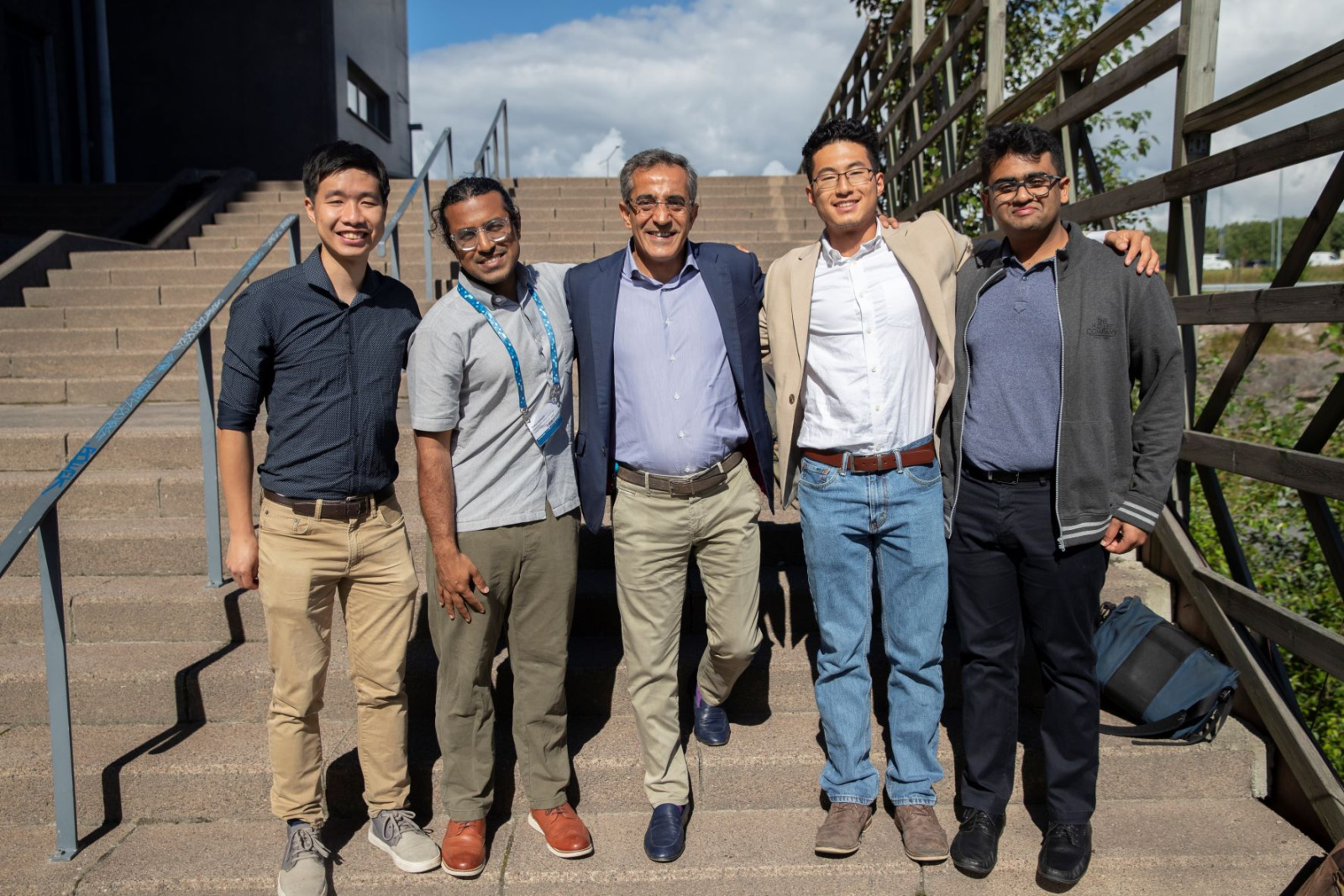 Five men stand arm and arm smiling at the camera standing on cement steps outside on a sunny day