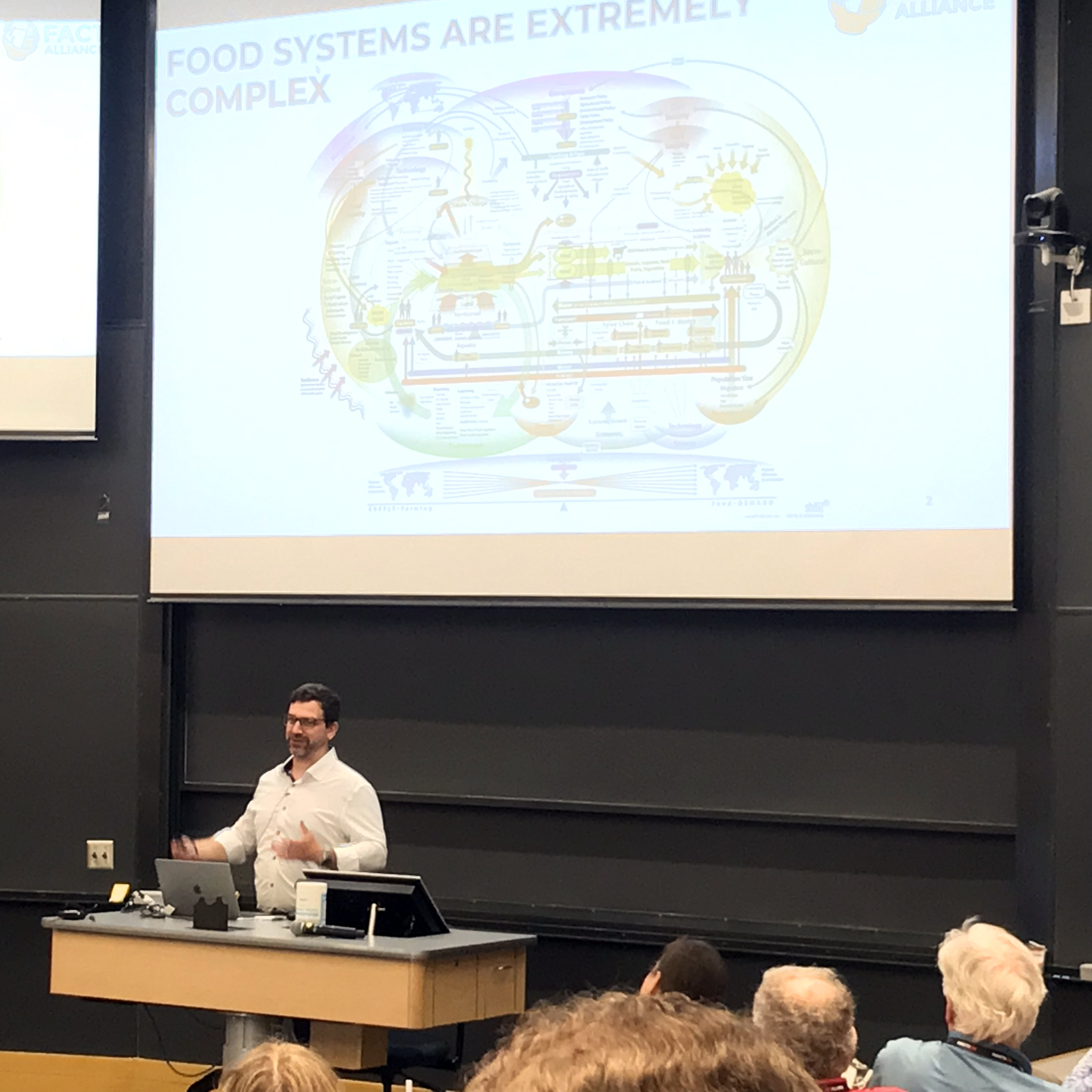 Greg Sixt presents in front of a large presentation screen to a group of people