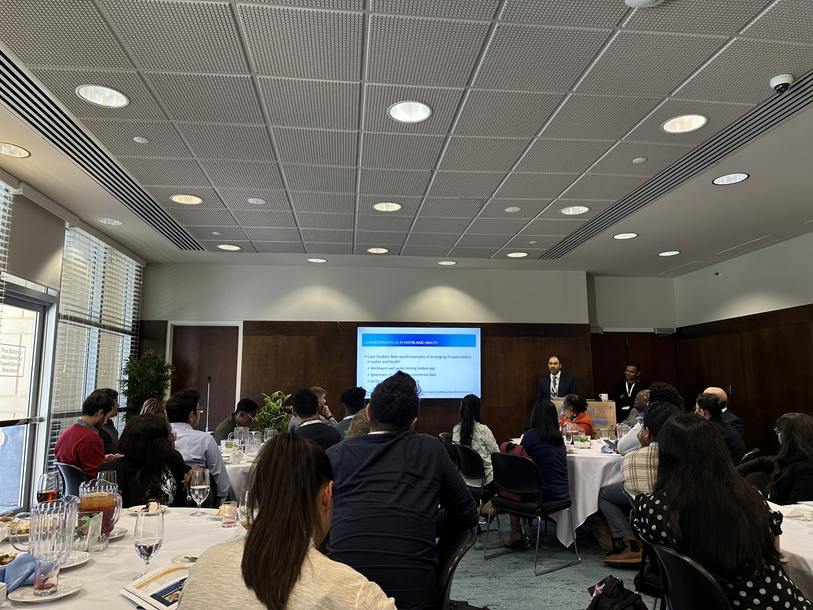 Conference attendees at round tables listening to session, screen reads as "Understanding AI in Water and Health."  