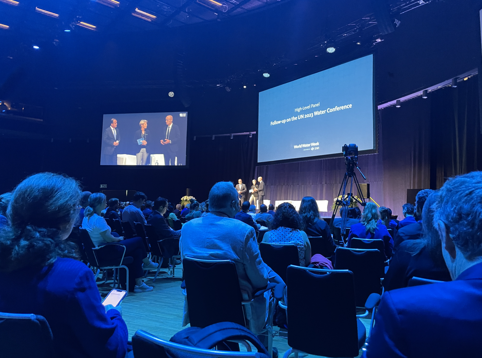 Three speakers at the front of a packed conference room beside a presentation slide displaying 'High-level Panel Follow-up on the UN 2023 World Water Conference.'