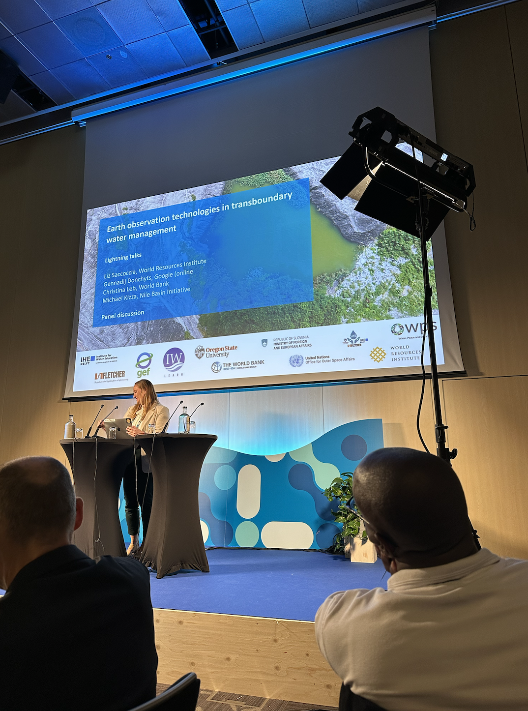 A speaker behind a podium addressing a crowd in from of a projector that says, "Earth observation technologies in transboundary water management."