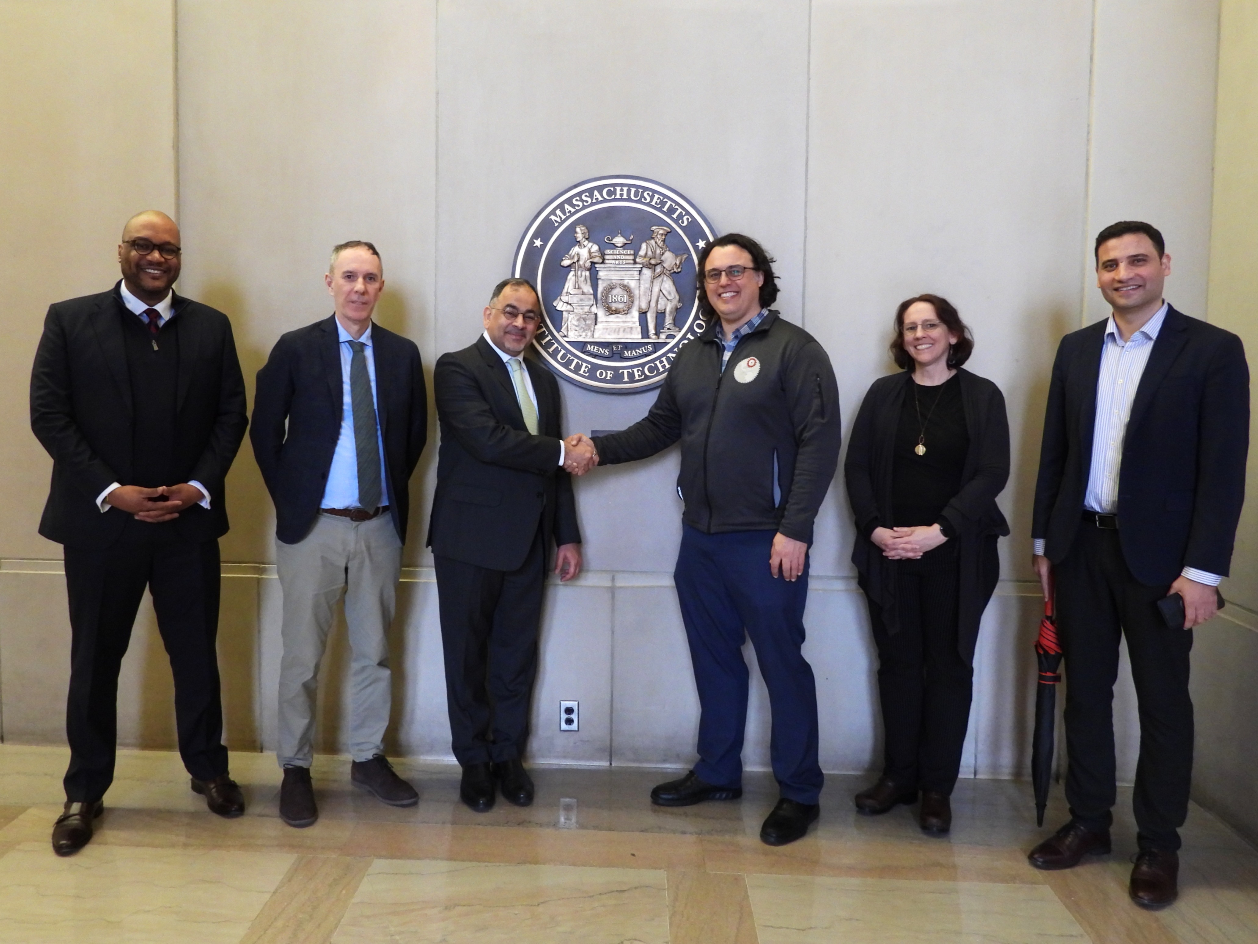 A group of six people stand in front of the MIT emblem on a wall. The two people in the middle are shaking hands