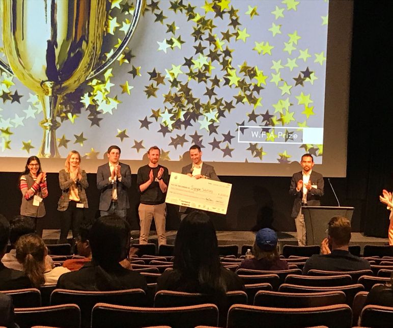 Award winners stand on a stage in an auditorium with one of the winners holding a check