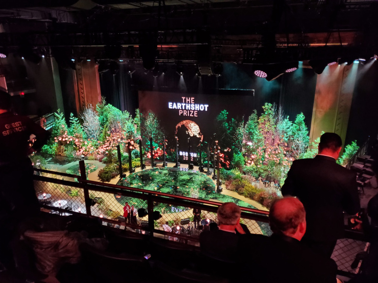 View of a large stage from a theater balcony, showing trees and shrubs and a large screen that says Earthshot Prize Boston 2022