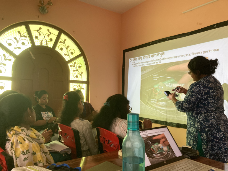 A teacher stands at the front of a room by a projection slide, demonstrating how to use the sensors to a group of people