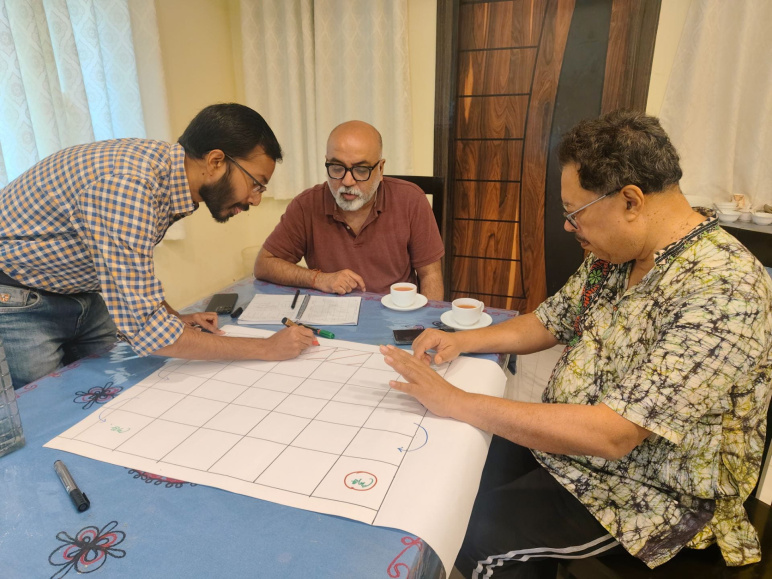 Anamitra Saha, Sai Ravela, and Jayanta Basu sit around a large poster sized paper at a table working