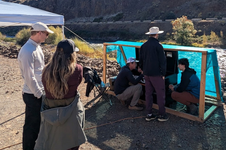 A group of people stand by a river, some are crouched under a tarp around a device that isn't visible