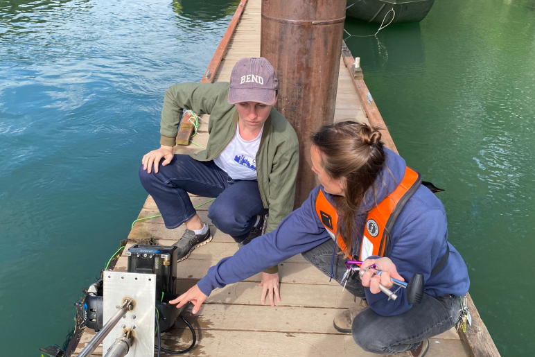 Professor Sara Beery discusses her sonar monitoring system with another researcher on a dock on a river..