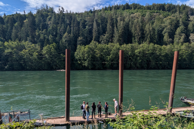 Six people in conversation stand on a dock next to a river with green trees and blue sky in the background
