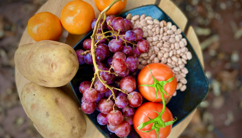 A table with a basket of food filled with tomatoes, oranges, potatoes, beans, and grapes