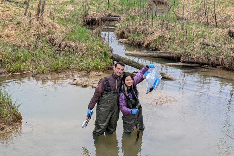 Two people stand in a swamp-like body of water wearing rubber overalls and holding up plastic bags with soil samples