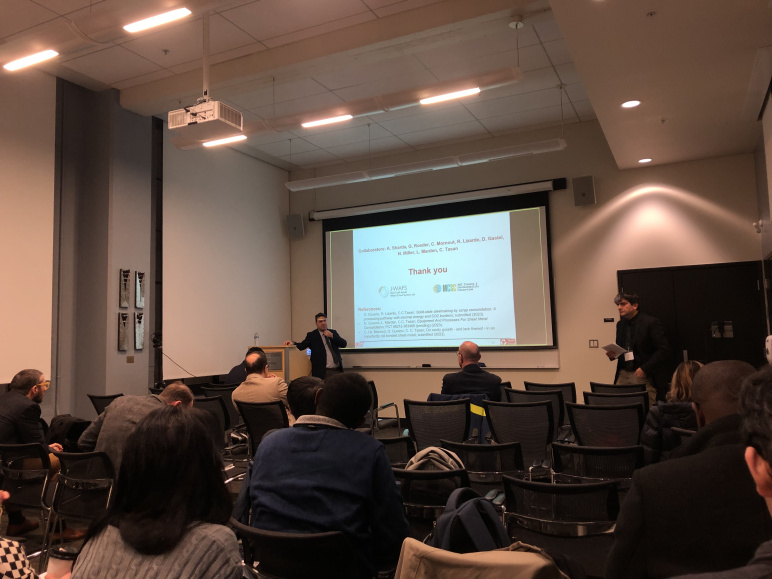 Onur Guvenc in a suit stands at the far left of a presentation screen and gives a presentation to a room full of people. Cem Tasan stands on the far right.