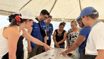 A group of people standing around a table,  pointing to and discussing the maps on the table