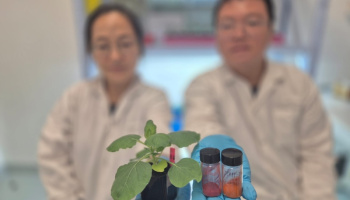 Two out-of-focus scientists in a lab. One holds up a small potted plant; the other, two small glass vials containing powder in different shades of red