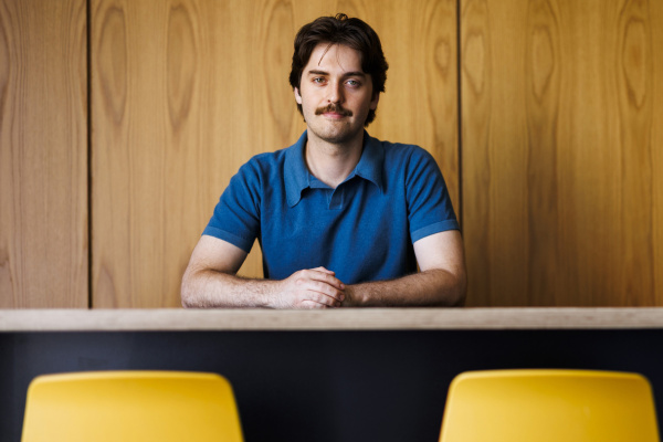 Coon posed sitting behind desk with hands crossed, light smile