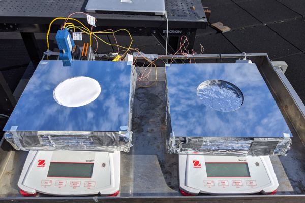 Roof top photo shows two scales, each holding thick square devices covered in foil. The blue sky is reflected.