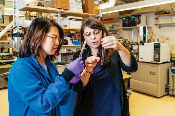 Desirée Plata, right, is wearing a blue lab coat and goggles while showing her grad student, left, a liquid in a test tube. This is taking place in a laboratory. 