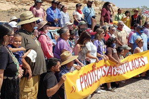 A group of around 50 people of all ages standing together outside in a desert-like climate holding a banner that says "Protect Acred Places" 