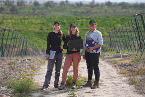 Three researchers from the GEAR Lab stand on a dirt road in a field in Jordan holding laptops.
