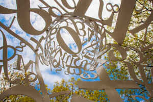 An artistic metal sculpture with intertwined numbers and letters, viewed from below against a backdrop of a clear blue sky and tree branches.