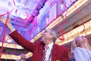 Older man and woman look up delightedly at red streamers falling from the ceiling.