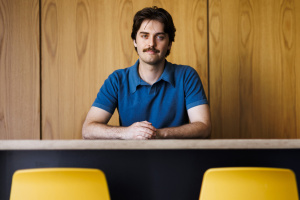 Coon posed sitting behind desk with hands crossed, light smile