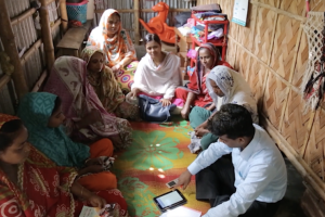 In Bangladesh, a group of eight women sit in a circle in a small room. A man, also sitting on the floor with them, is explaining a BRAC humanitarian program. An iPad and cell phone are on the floor. 
