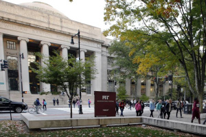 Dozens of people walk along the sidewalk and up the stairs of a large, ornate building at the MIT campus.