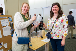 Photo of Theresa Werth and Dorothy Hanna standing at a table with Werth pouring a pitcher of water into a mug held by Hanna. A bulletin board next to them has pictures of natural water scenes and the words "I don't know where to fill my bottle?"