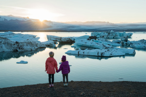 Two children holding hands looking at glaciers on the water with a sunset over the mountains in the background