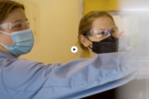 Ariel Furst and her student wearing masks, blue lab coats, and protective eye gear as they look at a white object that is blurry in the screenshot.