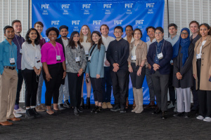 Photo of a large group of students standing together and posing in front of a blue banner with "MIT PKG Ideas" logos