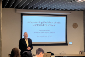 Dale Whittington giving a lecture with a presentation screen in back of him and people sitting at a conference table in front of him