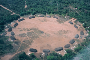 The village has about 20 huts that form a large a ring around an empty, brown, circular area. Lots of trees are around the village.