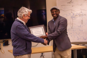 Two men are shaking hands and smiling as one hands the other a framed certificate, with a whiteboard containing diagrams and equations in the background.