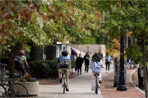 Three people ride a bike outdoors during a fall day at MIT campus with others walking in blurry background.