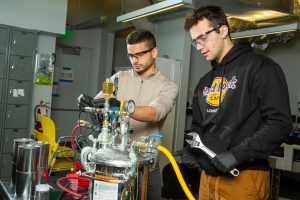 MIT engineers are in the lab wearing safety googles and working with tabletop device with many tubes and valves.