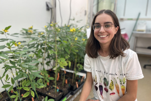 Photo of Sonia Boor sitting next to rows of plants in a lab greenhouse