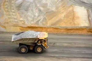 A large truck loaded with rocks drives along a road in a mining site, with terraced layers of excavated earth in the background.