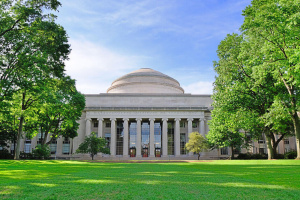 The front of the MIT main building with its large dome, seen from a grassy area with trees on a clear day.