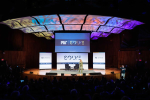 A speaker presents on stage at the MIT Solve event in an auditorium with colorful lighting and a digitally displayed backdrop.