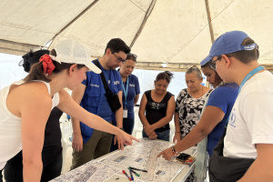 A group of people standing around a table,  pointing to and discussing the maps on the table