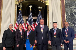 Six individuals standing in a line wearing medals; there are three American flags in the background. 