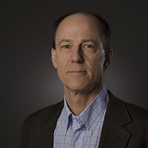Gregory C. Rutledge sitting with a blue shirt and gray jacket against a gray backdrop