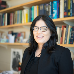 Tavneet Suri in a black blazer and glasses smiling in front of a bookshelf full of books