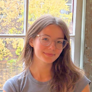 A headshot of Avery sitting in front of a window with fall foliage peaking through.