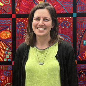 Daniela Giardina stands smiling in front of a colorful quilted tapestry of faces